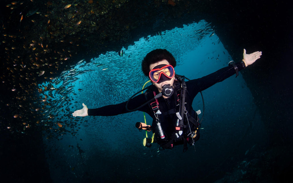 A scuba diver with arms outstretched underwater, surrounded by a school of fish near a submerged structure.