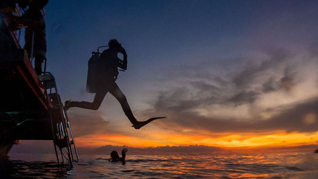 a scuba diver stepping off a platform into the water while the sun sets in the background